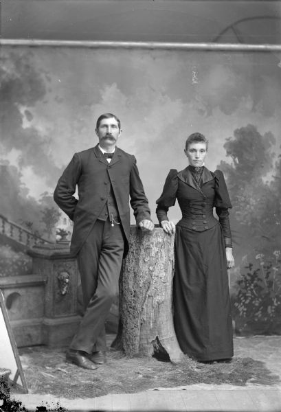 Studio Portrait of European American Couple Full-length studio portrait of a man posing standing on the left and a woman posing standing on the right, with their hands resting on a stump in the center.