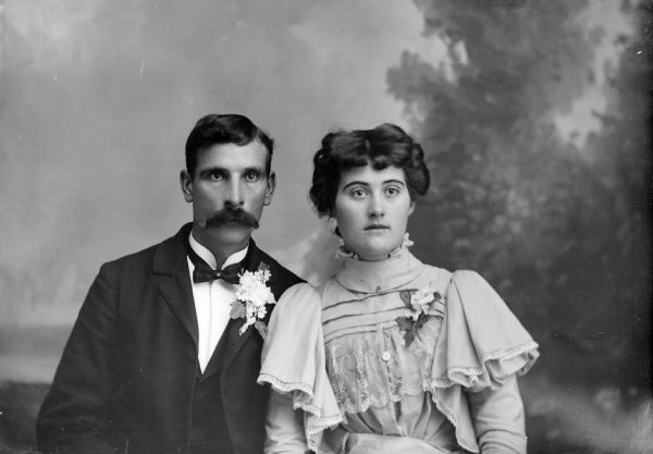 Studio Portrait of Unidentified European American Couple Waist-up studio portrait in front of a painted backdrop of a man and woman posing sitting. They are both wearing corsages.