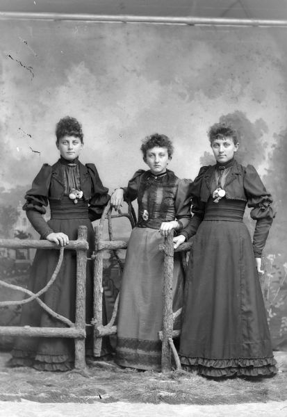 Group Portrait of Three Unidentified European American Women Full-length studio portrait in front of a painted backdrop of three woman posing standing at a prop wooden fence and gate. All three women are wearing flowers at their waists.