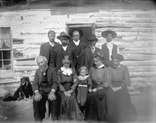 Outdoor Group Portrait of European American Family Outdoor group portrait of a large unidentified family group posing sitting and standing. Five men are standing behind three women, a man and a young girl sitting on a bench. A dog is sitting next to the log building in the background on the left.