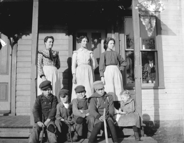 Outdoor Group Portrait of European Americans Outdoor portrait of an unidentified group of people posing on a front porch. Three women are on porch, and four boys and one girl are sitting on the porch steps. The girl is holding what may be a dog sitting behind her and one of the boys. Two other people are standing inside the house looking out of the window on the right.