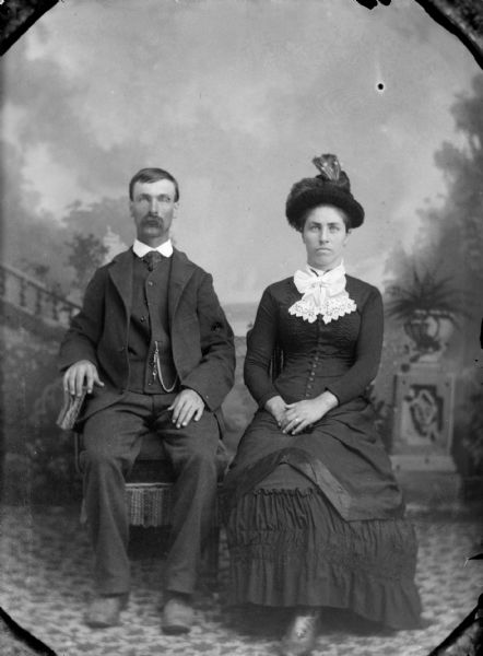 Studio Portrait of European American Couple Full-length studio portrait in front of a painted backdrop of an unidentified man and woman posing sitting. The man is wearing a dark-colored suit coat, trousers, vest, necktie, and watch chain. The woman is wearing a dark-colored dress with a button-down bodice and a white lace collar.