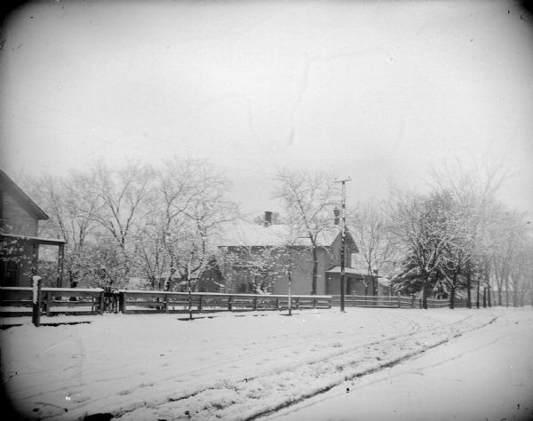 Snowy Scene on Fifth and Main Streets Outdoor view of a snow-covered street and houses behind a long wooden fence. Location identified as the intersection of Fifth and Main Streets looking west.