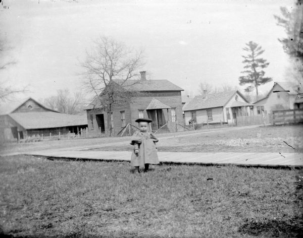 Girl Next to Plank Sidewalk Outdoor portrait of a small European American girl posing standing near a wooden walkway in a yard in front of several buildings.
