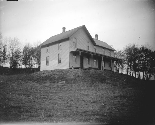 House on a Hill Outdoor portrait of a two-story wooden house on a grass-covered hill with a line of trees behind it.