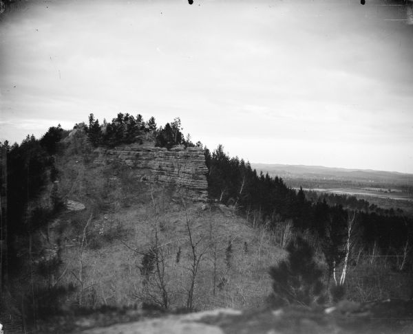 Castle Mound View from hill towards a rock formation rising up from a slope and surrounded by trees. Location identified as Castle Mound. In the background on the right are trees and fields, and low hills on the horizon.