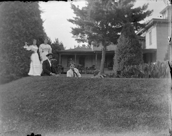 The Spaulding Family at Home Outdoor group portrait of a man and two women posing sitting on the lawn just behind a low hill, and two women posing standing behind them near tall bushes, in the yard of a large house in the background. Identified as the Spaulding family in the yard of their residence.