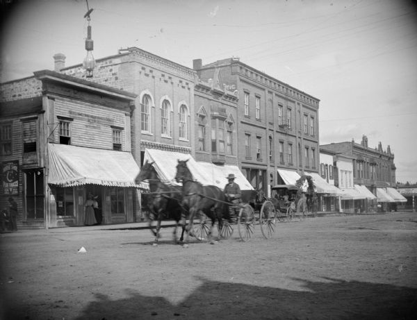 Scene on Main Street Outdoor view from side of street towards a man driving a wagon moving down a town street pulled by a team of two horses. Location identified as Main Street.