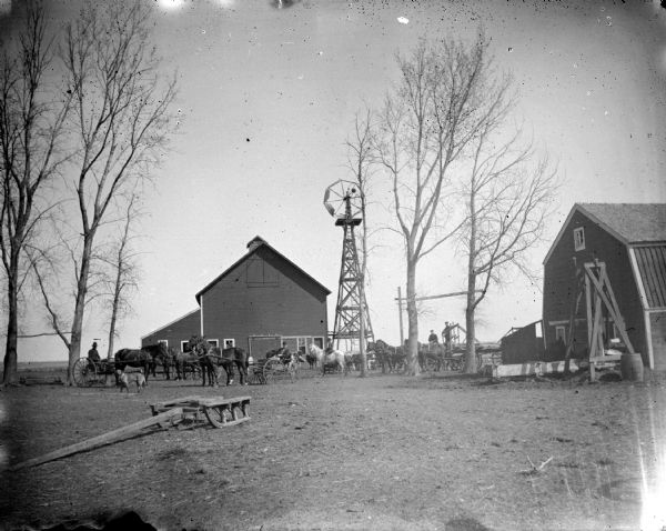 Farm Scene View across barnyard towards men sitting in several wagons and buggies. Around them is a tall windmill and two barns.
