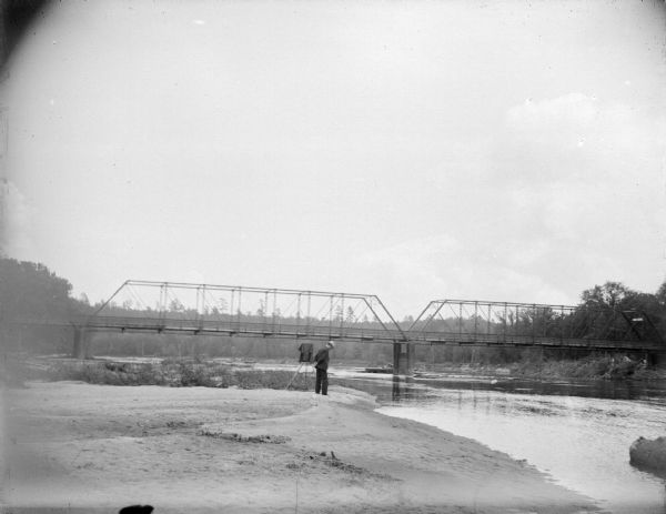 Man Near Melrose Bridge View along shoreline towards a man posing standing on the sand, with what may be a camera on a tripod. In the background is a bridge. Identified as probably the bridge across the Black River at Melrose.