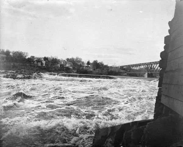 Black River Scene View towards the shore of a river with a wall or foundation in the right foreground. On the opposite shoreline are buildings, and a bridge over the river on the right.