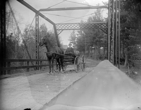 Man in Horse-Drawn Buggy on Bridge View along railing of bridge towards a horse pulling a man in a buggy on a bridge. The unpaved road in the background has trees on both sides, and there is a building in the distance.