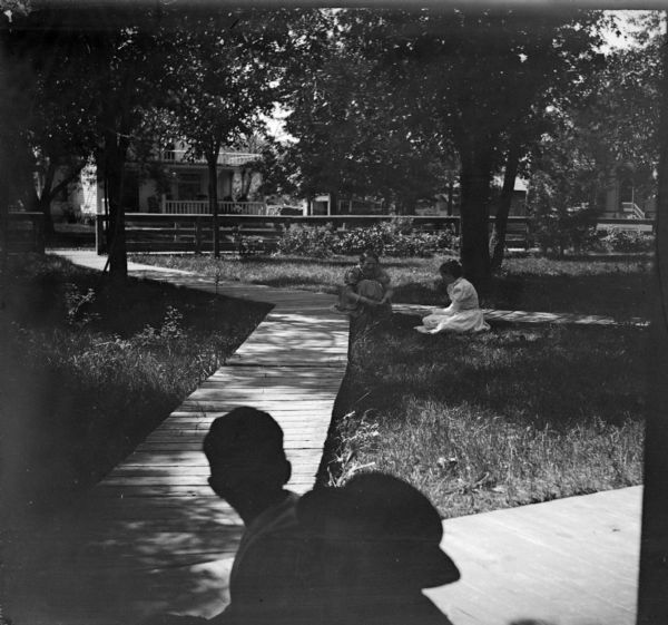 Four People Sitting in a Park View towards two women posing sitting on the ground at the intersection of two wooden walkways in a park. Two men, in shadow, are sitting in the foreground.