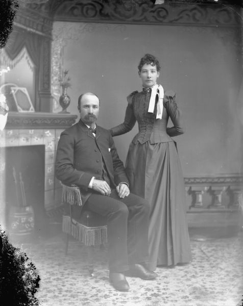 Studio Portrait of Unidentified European American Couple Studio portrait of a man posing sitting on the left and a woman posing standing on the right. The woman has her hand on the back of the chair.