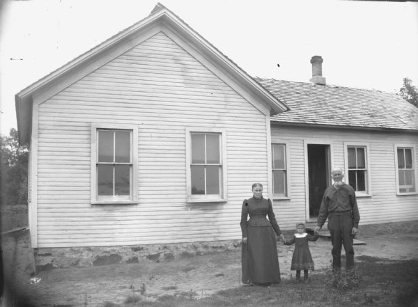 European American Family Posing in front of their Home View across lawn in an unidentified location of an elderly woman and elderly man posing standing and holding the hands of a girl posing standing between them. They are in front of a white clapboard house.
