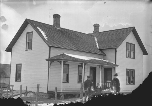 Group Portrait of European American People in front of House View towards a man, woman, and two children in an unidentified location. The children are standing between the man and woman, and in the background is a two-story wooden house. Snow is on the ground.