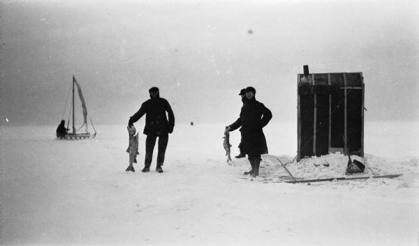 Two men holding large fish pose near an ice fishing shanty off of Fish Creek. Another man stands behind them. There is man on an iceboat in the left background.