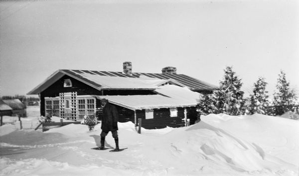 Ferdinand Hotz poses wearing snowshoes at his cottage in Fish Creek, now 4108 Main Street. There is a large snowdrift in the foreground.