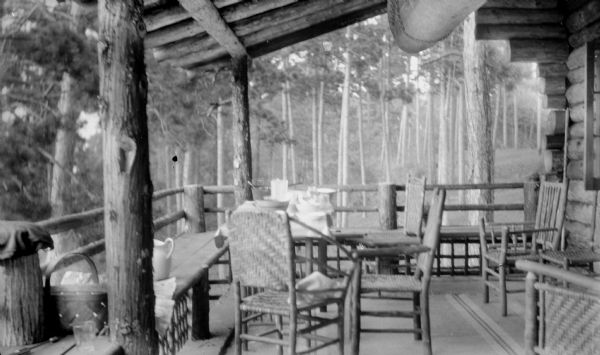The porch of the Hotz cottage at Europe Lake, furnished with a rustic table, chairs and benches; the table has been set for a meal. The cottage and porch are of log construction.