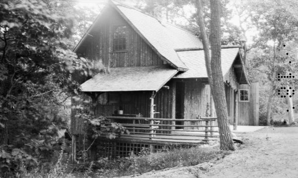 A vertical log-sided garage with lean-to porch stands on the side of a hill in the woods. There is a rustic table on the porch.  This building was located across the drive from the Hotz cottage.