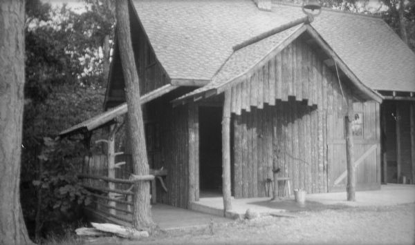 The garage, with vertical log siding, near the Hotz family cottage at Europe Lake. There is a pump on the front porch, and a bell with a rope on the peak of the roof. There is a lean-to porch on the side.