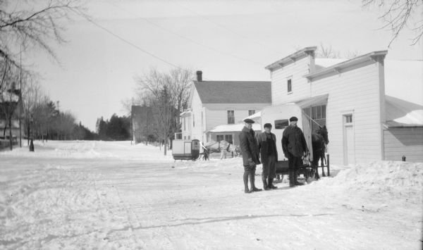 Three men stand behind a horse-drawn enclosed sleigh in front of the Lundberg Store on Fish Creek's Main Street. There is a second sleigh and horse in the background in front of the Hotel Barringer (Central Hotel).  Dr. Welcker's Casino is nearly obscured by trees beyond the hotel.