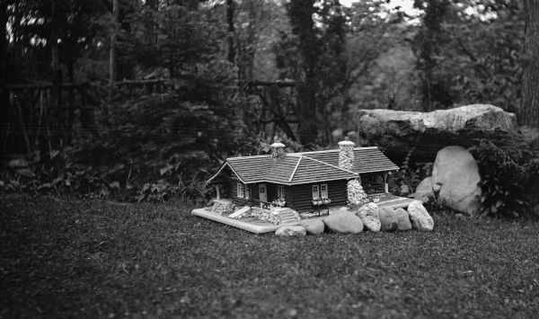 An architectural model of a log cabin with rustic details and stone chimneys rests on a lawn. There are trees and a rustic railing in the background.  The model is of the Hotz cottage at Europe Lake in Door County, WI.
