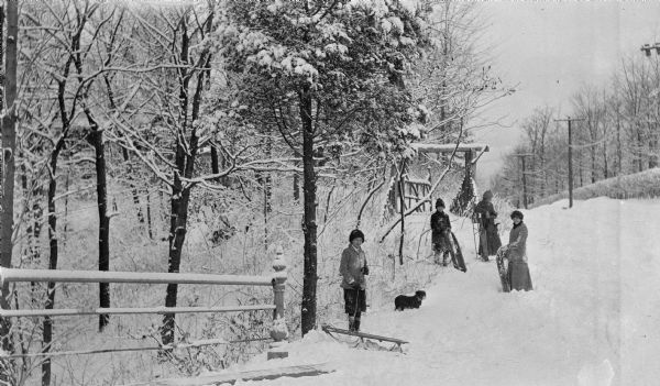 Four children pose in the snow with their sleds and a dachshund. There is a rustic bridge in the background and a railing in the left foreground.