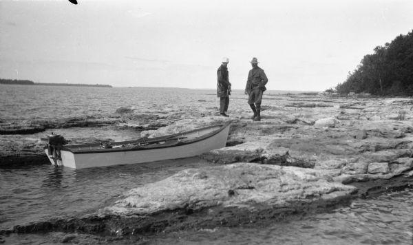 Two men, including Ferdinand L. (Fedy) Hotz, left, stand on the west shore of Spider Island off Door County.  There is a small boat with an outboard motor in the foreground.