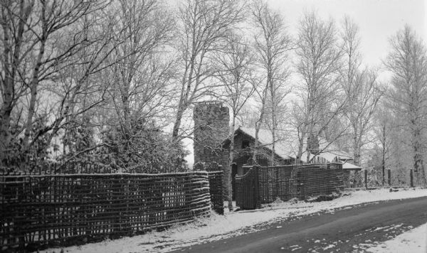 View of the stone garage and tower at the Hotz Fish Creek compound on Egg Harbor Road, now Highway 42. There is a rustic lattice fence and gate along the road. Light snow covers the birch and evergreen trees.