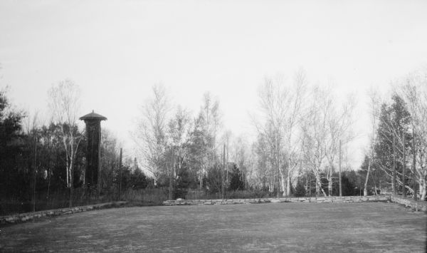 The tennis court at the Hotz Fish Creek compound is surrounded by a low stone wall and tall posts. Outside the wall at the far left corner of the lawn is a tall wooden tower with a roof. The stone garage and tower are faintly visible through the trees in the background.