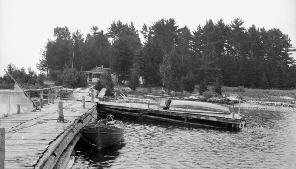 A boat with an outboard motor is tied to the rustic log pier; a second boat is overturned on an extension of the pier. There is a fish net hanging over the railing. In the background is a small cottage with a massive stone chimney and two screened porches.  This cottage on North Bay was built by Sturgeon Bay dentist Arthur J. Gordon, D.D.S. and his wife Maude.