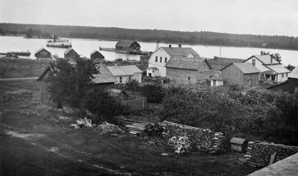 Elevated view of Fish Creek Harbor as a boat approaches the pier and warehouse. In the foreground are houses with outbuildings, gardens and a small orchard. Cordword is stacked along a fence.
