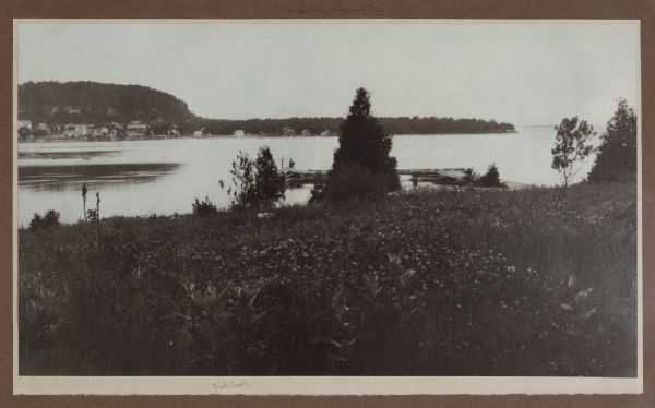 View down hill towards a man fishing from the pier at Nelson Point in Peninsula State Park. Fish Creek is seen across the bay. Dr. Welcker's Casino is the large building with gabled roof and porch at the base of the bluff. An outbuilding in the center is labeled "Welcker's Laundry."