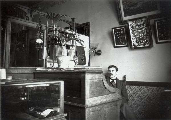 A young Ferdinand Hotz sits behind a large desk in his Chicago office. There is an art print and a framed collection of moths and butterflies on the wall behind him. There is a nearly empty glass display case in the foreground.