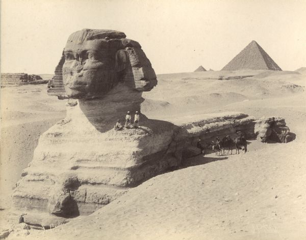 The Sphinx at Giza in Egypt. Three men are standing or sitting on the shoulder of the Sphinx, and several men are sitting in the shade below with two camels, and one man astride a donkey. Pyramids are in the background.