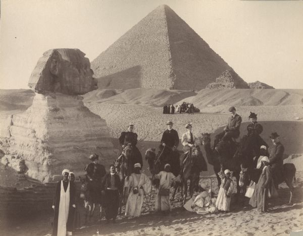 A group portrait of Violet Lee Dousman (back row, 3rd from left) and her fellow travelers, guides and Egyptians on a trip to Giza, Egypt. Five people are riding camels and one man and one woman are mounted on donkeys. Behind them are the pyramids and another group of travelers, on the left is the Sphinx.