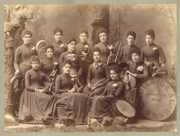 Studio portrait in front of a painted backdrop of the Ladies Ideal Band, in uniform and holding their instruments which include drums and various brass instruments. Text from an article in the Mauston Star, July 4th, 1946, "Band Belles of the 80's - The members of Mauston's famous Ladies Ideal Band included: Front Row (left to right) Alice Stewart, Ella Schall Van Wie, and Jessie Grimmer Heath. Second Row: Inez Wetherby, Emily Bury Earle, Adelaide Parker Winsor and Dora Wells Ramsey. Third Row: Nora Hall Dixon, Emma Priest Hinton, Nettie Priest Lyon, Grace Baldwin Price, Rosabelle Carter Winchell and Bertha Stewart. Unfortunately Jessie Hinton Bunnell and Emma Anderson Bradley, a founder of the band, as well as Mattie Sykes Bowes, and Agnes Wetherby Boorman are not shown in the picture. Although only Mrs. Bradley was married at the time, married names are given, where it is appropriate, to avoid confusion."