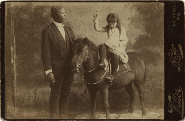 Cabinet card of an African American man, wearing a formal suit, standing next to a young girl mounted side saddle on a pony. She is reading a book and has her hand raised, holding a pen or pencil. She is wearing a dress with dark stockings and shoes. The image was taken in a photographic studio with a painted backdrop. The pony may or may not be stuffed.