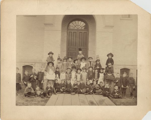 First and second grade children with their teacher, Miss Drinker, who was temporarily taking charge of Miss Ella Larkin's school. Taken in the front entrance to the old high school at the corner of Wisconsin Avenue and West Johnson Street. The children were taught in the room at the northwest corner of the building for lack of a better location. Boys: Charles Abbott, Paul Fish, Arthur Olson, Everette Green, Tony Brown, Jack Venetta, ? McKay, and Fred Chamberlin. Girls: Ada Welch, Ruby Peck (Mrs. John Corscot), Cora Norsman (Mrs. R. Neckerman), Thusuelda Helm (Mrs. A. Frautchi), Bettina Jackson, Gertrude Higham, Susie Nelson, Elsie Bodenius, Florence Livermoor, Mrs. Elsa Suhr, and Laura Janeck.