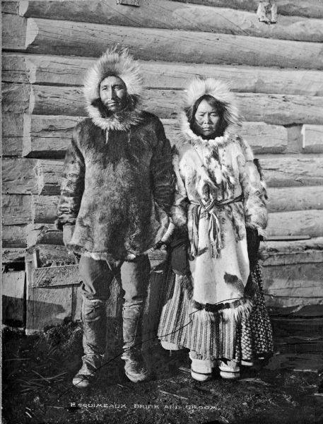 Full-length outdoor portrait of an Esquimeaux (Eskimo) bride and groom. They are wearing native clothing made from cloth and animal skins and are standing in front of a log cabin. Caption reads: "Esquimeaux (Eskimo) Bride and Groom."