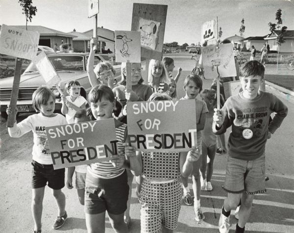 A group of children are holding up hand-made signs, demonstrating for "Snoopy for President." Several children are wearing Peanuts t-shirts and sweatshirts, and they are walking down the middle of a residential street.