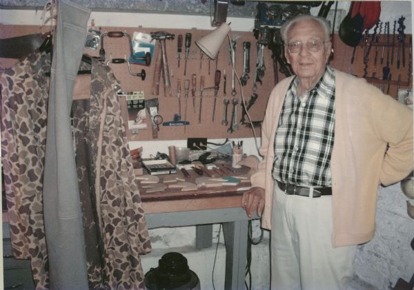 Fred O. Petzold (1911-2011) of Oshkosh, Wisconsin, in a cardigan sweater and plaid shirt stands next to his workbench. His camouflage hunting clothing is hanging on the left. Tools are hanging on a pegboard attached to the wall. Pieces of duck decoys appear on the top of the workbench, next to carving tools. A shop vac can be seen under the workbench.