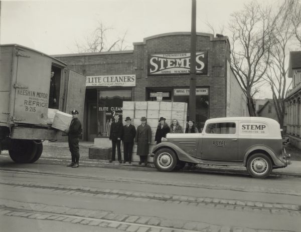 Delivery for Stemp Typewriter Company View from across State Street of men and women standing in front of the Stemp Typewriter Company as deliveries are stacked on the sidewalk. Stanley P. Stemp (owner) is fourth from the left and his wife, Elisabeth, is standing next to him. A Stemp delivery truck is parked to the right and a large delivery truck is on the left. A deliveryman is unloading boxes. The business on the left is Elite Cleaners, "For Particular People."