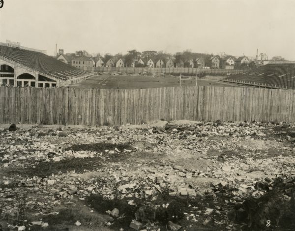 View of football field and bleachers from railroad embankment. In the foreground is rubble against a wooden fence. In the middle of the football field a number of men are working with shovels. A wooden fence on the far side separates it from houses in the neighborhood. Numerous piles of lumber are all along the foot of the fence in the field and behind the bleachers, and a scoreboard and ladder are against the fence. Text at foot of photograph reads, "Park Hill Place 9+69 Looking North from Line."