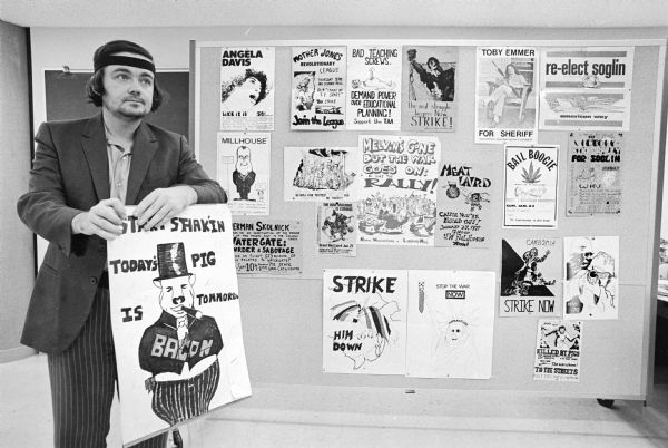 J. Wesley Miller posing with some of the campus street posters and handouts he collected. Mayor Soglin, Toby Emmer, Richard Nixon, Melvin Laird and Angela Davis are a few of the people portrayed in the posters. In 1973, he handed over 17,000 artifacts, including buttons, handouts, picket signs, posters and photos of messages sprayed on campus walls to the Wisconsin Historical Society. The collection was dated from the Fall War Moratorium, September 1970 to Election Day, 1972. Today the collection is known as "Madison People's Poster and Propaganda Collection." He collected the items on his way to and from classes at the University of Wisconsin. He dated each piece on the back giving Wisconsin a complete "representative history" of the University of Wisconsin-Madison's most turbulent times.