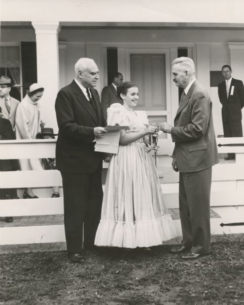 Ruth DeYoung Kohler II, presents the key to Wade House to Dr. William McKern, of the Wisconsin State Historical Society. The century-old stagecoach inn and adjoining buildings were purchased by the Kohler Foundation, Inc., in 1950, and restored under the direction of the young lady's mother, the late Mrs. Herbert V. Kohler. Her father, Herbert V. Kohler, chairman of the Kohler Foundation, is standing on the left. Other people can be seen standing behind the white fence and on the porch.