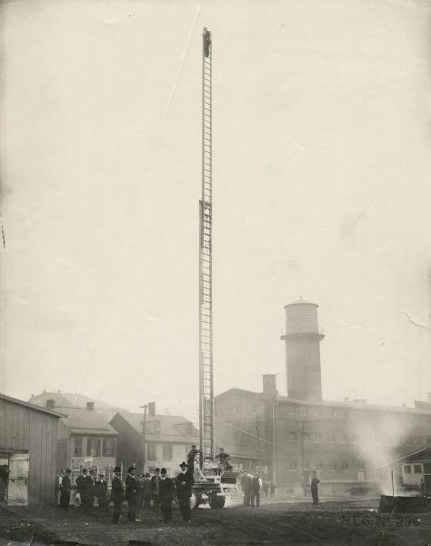 Fire fighting scene in which a man is at the top of a very tall ladder mounted on a hook and ladder fire truck. A group of men observe. A haze of smoke fills the air. Factory buildings and two houses are in the background.