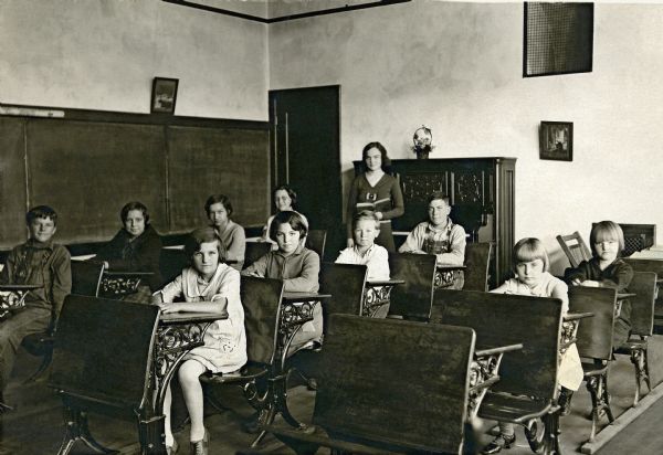 Ten students sitting at their desks in the classroom at Bay Hill School. Their teacher, Alice Holloway, stands at the back holding a book. Blackboards cover the wall on the left. A door and a piano are along the back wall, on which hang two framed images.