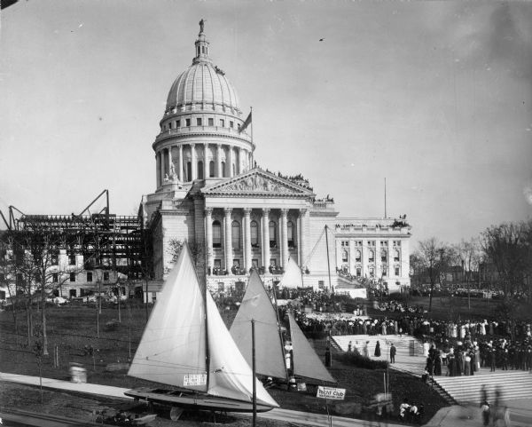 Elevated view of the Wisconsin State Capitol building, showing steel work of North Wing under construction. Crowds of people are gathered on the lawn for the Fall Festival. More people are gathered on the roof of the Capitol building just below the dome. Two boats are on display in the foreground near a sign that reads: "Mendota Yacht Club 'Boost Madison Lakes.'" The iceboat on the right is the <i>Princess II</i> and the sailboat on the left is the <i>Neireid</i>.
